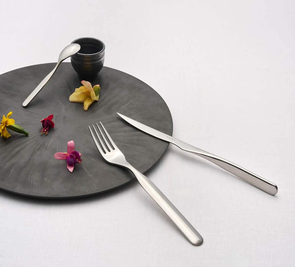 Cubertería Bugatti en acero inoxidable sobre plato de pizarra con flores comestibles – menaje para hostelería