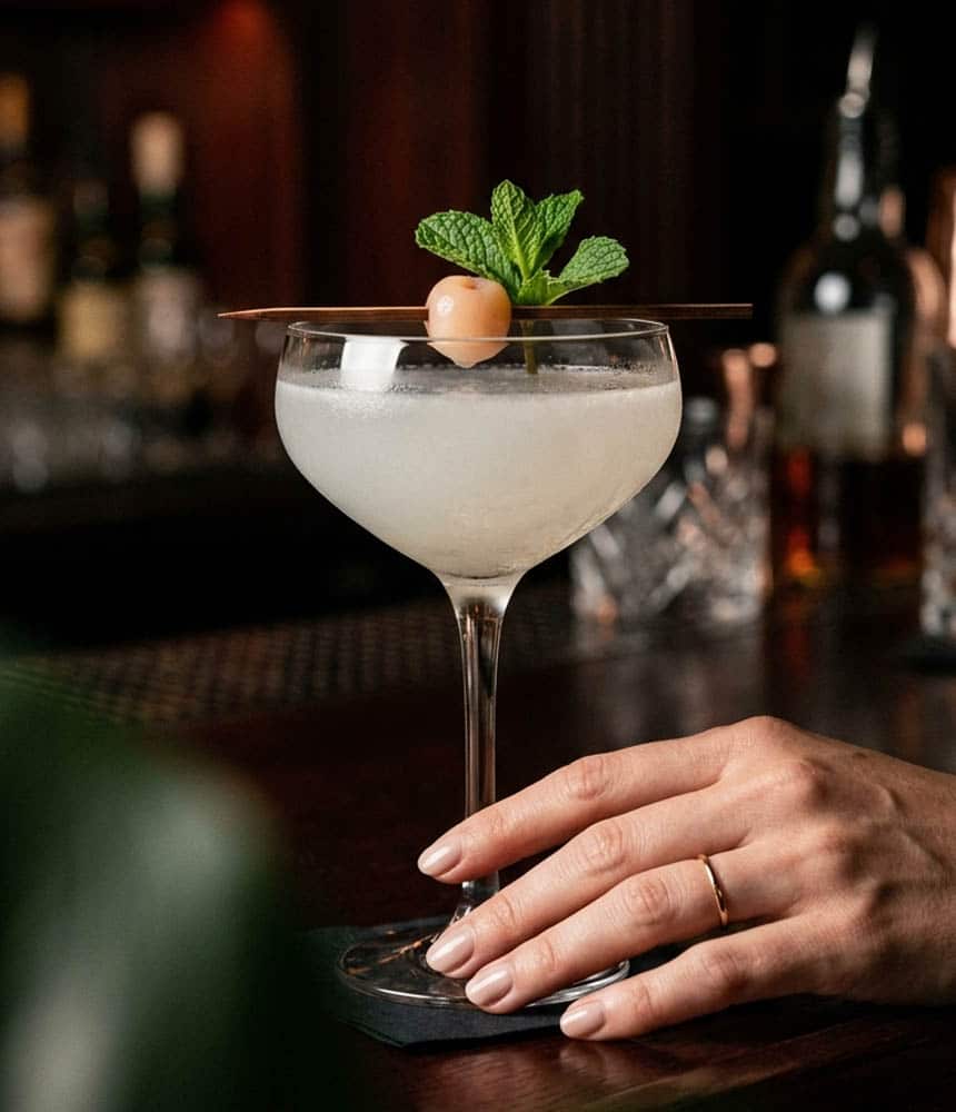 Copa coupe con un cóctel claro y garnish de menta y lichi sobre la barra de un bar