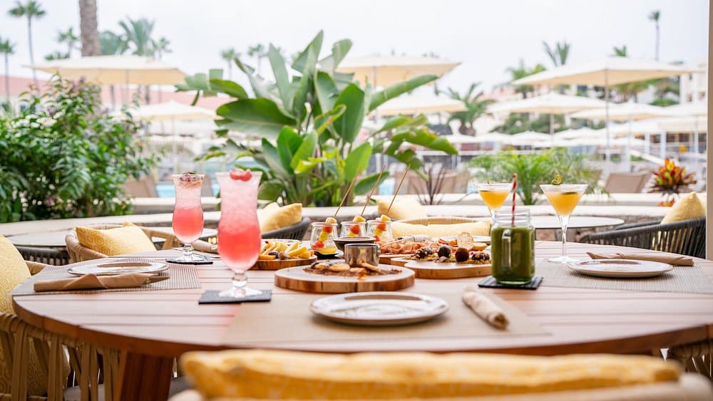 Mesa de terraza de hotel con cócteles, copas de martini, tablas de picoteo y sillas de ratán junto a la piscina