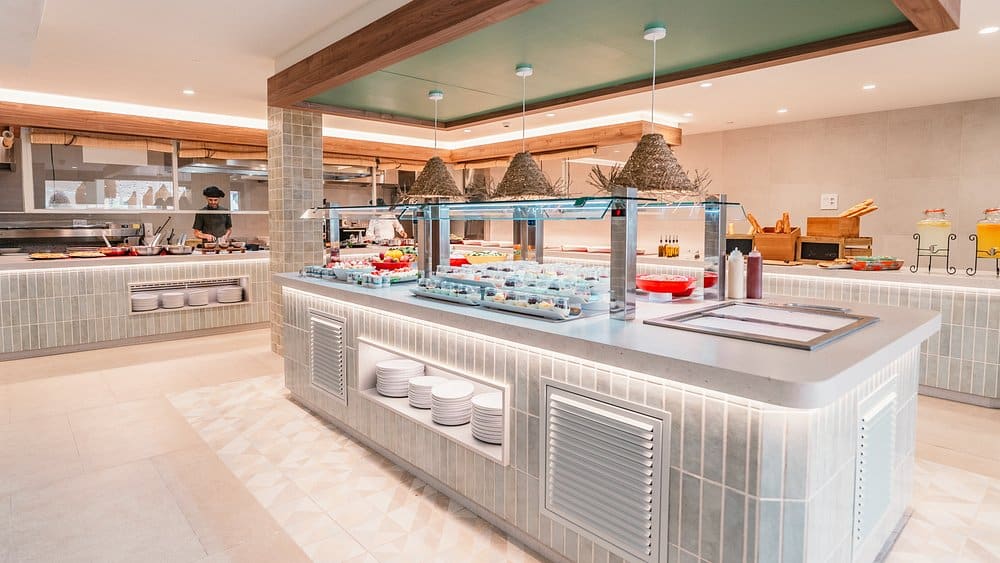 Isla de buffet de hotel con vitrina fría de raciones individuales, platos apilados en hornacinas y zona de cocina en vivo al fondo